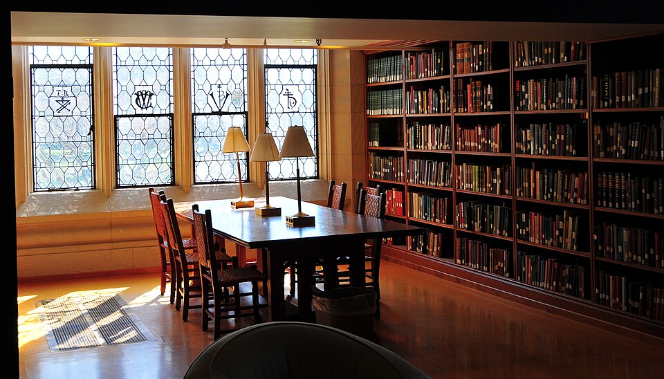 A wood-paneled library reading room with a long table, brass lamps, shelves of old books, and leaded-glass windows